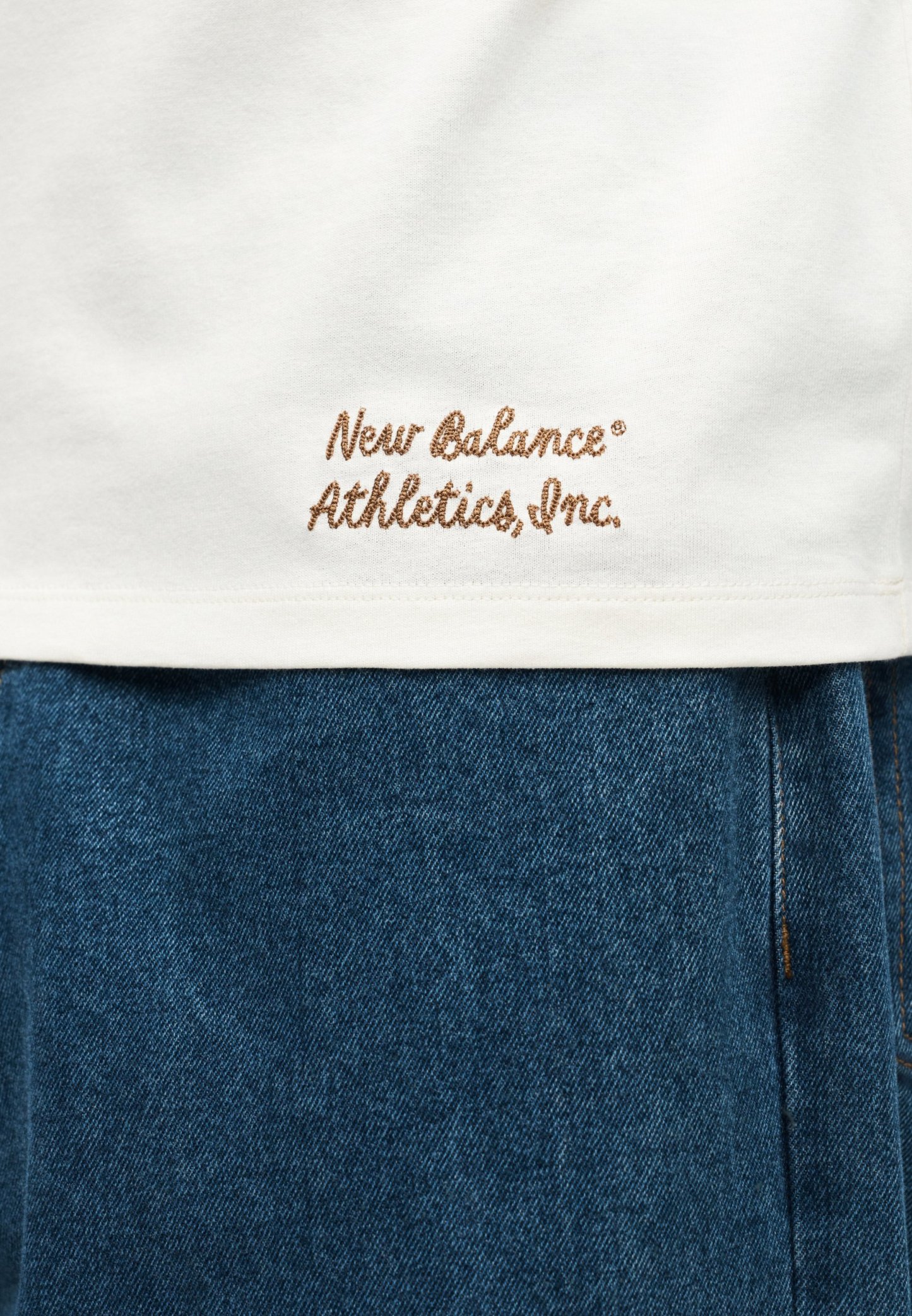Dobladillo de camisa blanca con bordado marrón "New Balance Athletics, Inc." sobre pantalones vaqueros azules de denim.