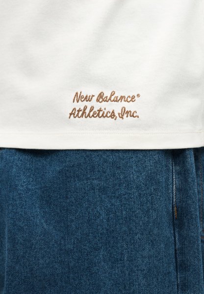 Dobladillo de camisa blanca con bordado marrón "New Balance Athletics, Inc." sobre pantalones vaqueros azules de denim.