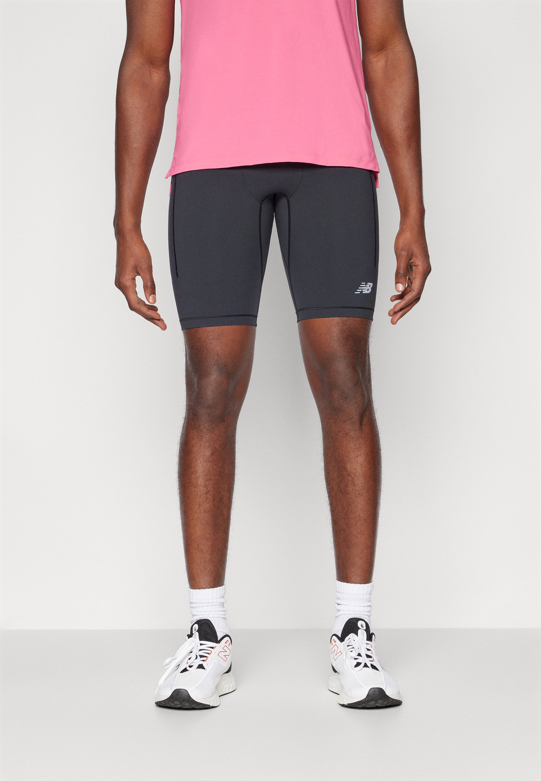 Shorts de compresión negros con un diseño ajustado y un logo reflectante de New Balance. Llevados con una camiseta deportiva rosa y zapatillas blancas con acentos.