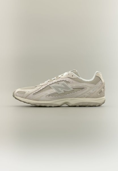 Zapatilla deportiva New Balance beige con paneles de malla y ante, cierre de cordones y suela acolchada, mostrada de perfil sobre un fondo neutro.
