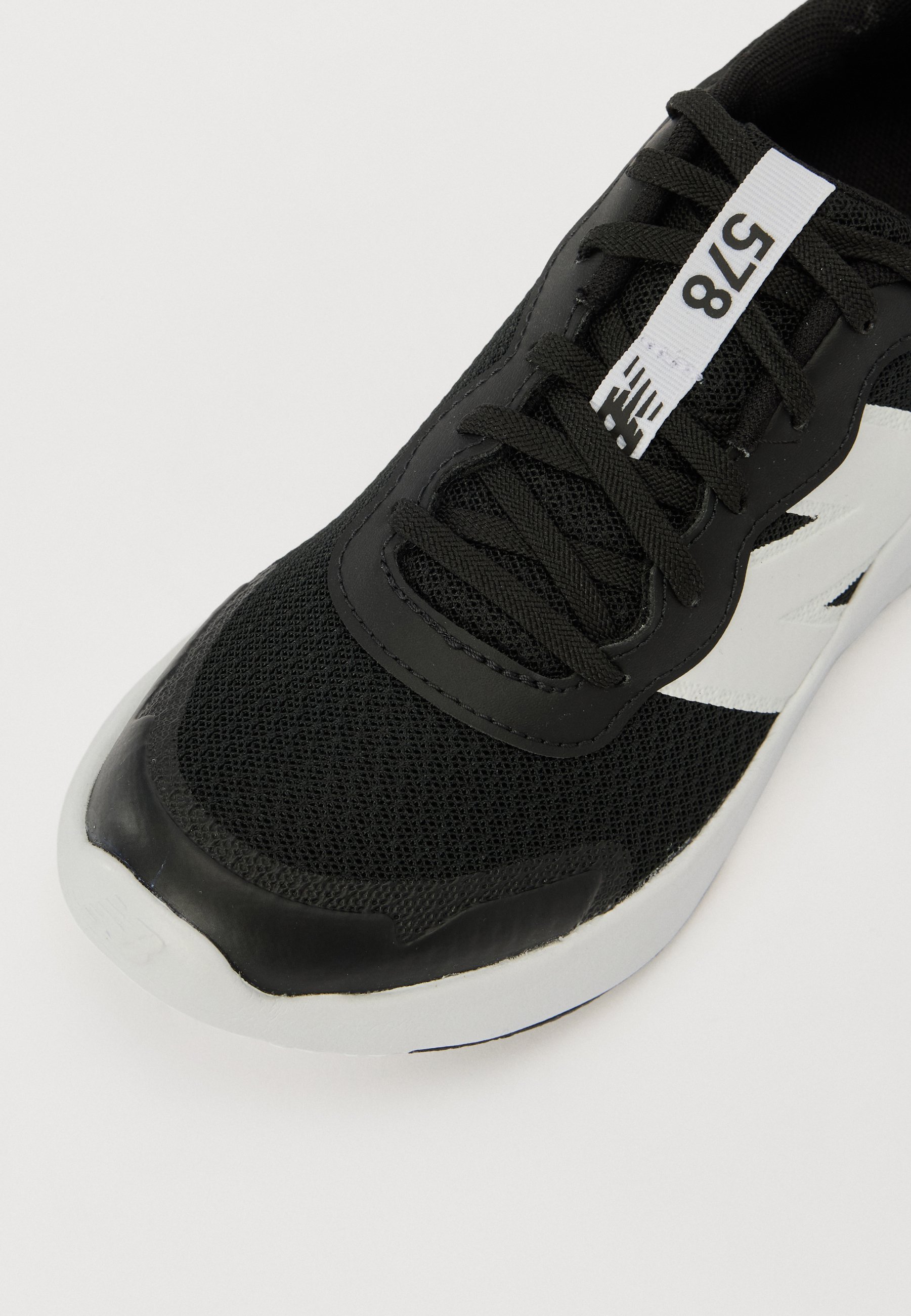 Zapatilla deportiva negra con parte superior de malla, puntera de goma y detalles en blanco. Presenta cordones y una etiqueta con números en la lengüeta.