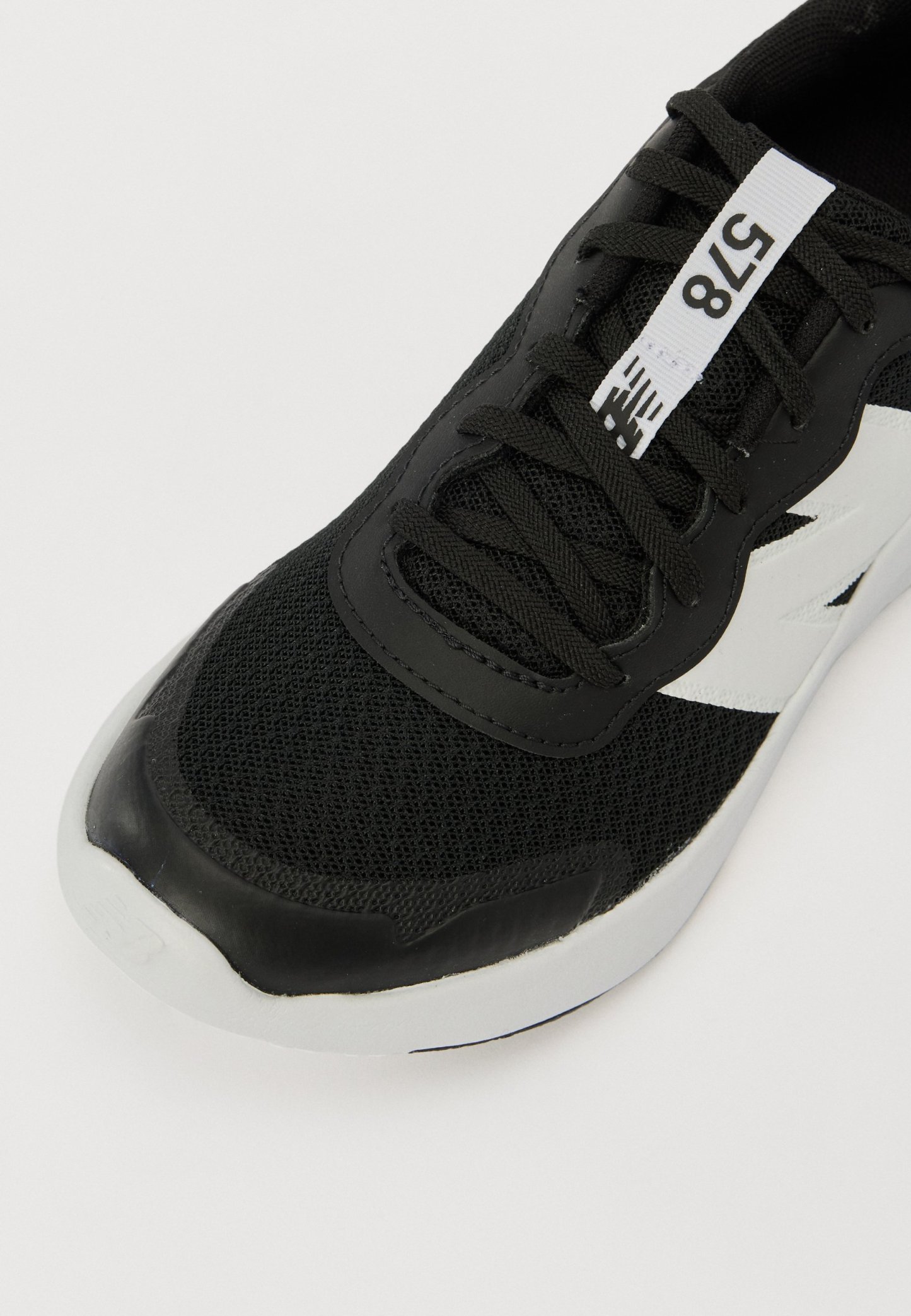 Zapatilla deportiva negra con parte superior de malla, puntera de goma y detalles en blanco. Presenta cordones y una etiqueta con números en la lengüeta.