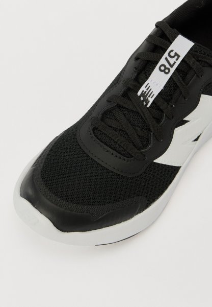 Zapatilla deportiva negra con parte superior de malla, puntera de goma y detalles en blanco. Presenta cordones y una etiqueta con números en la lengüeta.