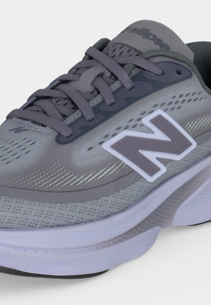 Zapatilla deportiva New Balance gris con suela blanca, parte superior de malla y cordones grises, con un gran logo "N" blanco y gris en el lado.