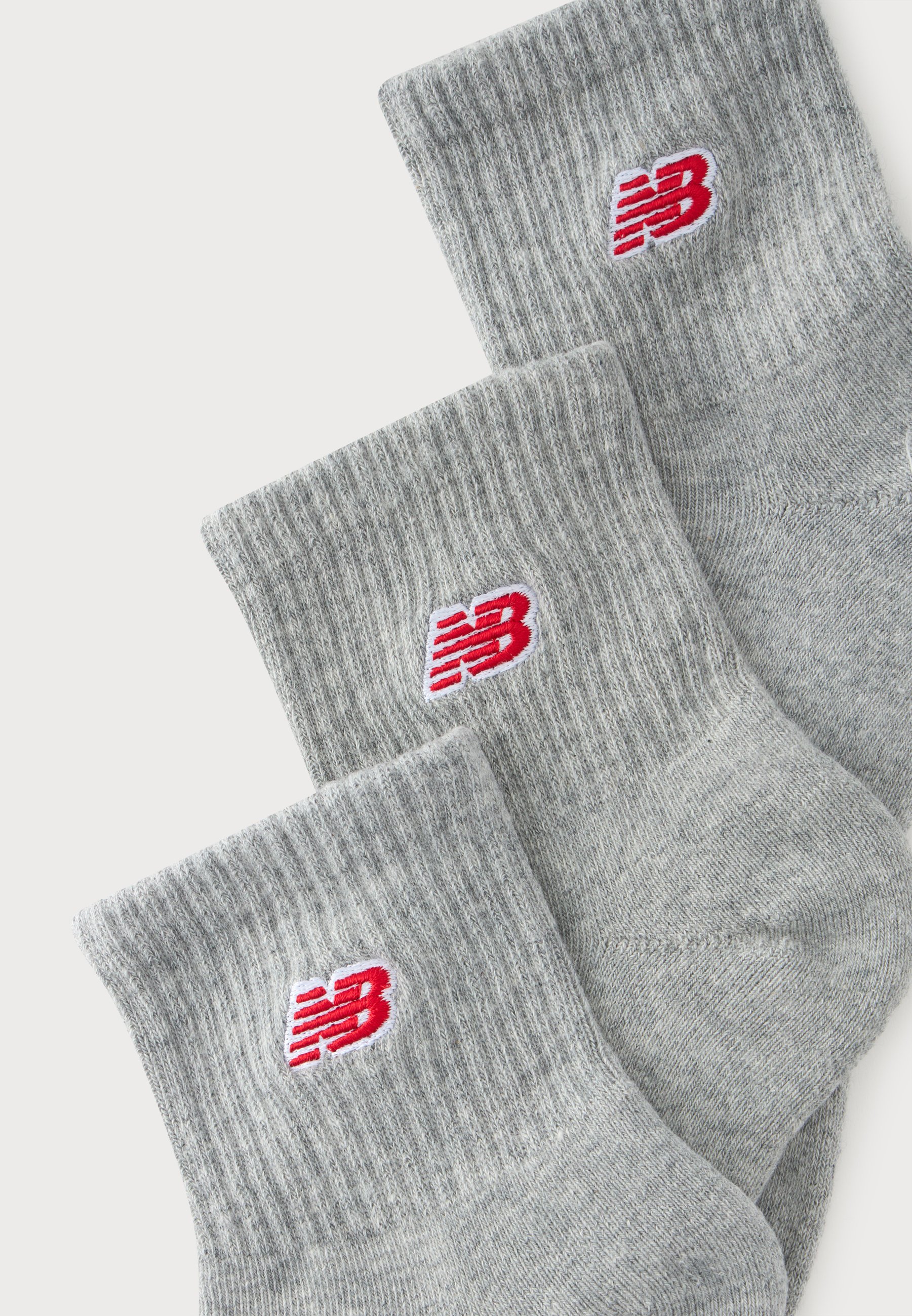 Tres pares de calcetines grises acanalados con logotipos rojos de New Balance en los puños, dispuestos superpuestos sobre un fondo blanco.