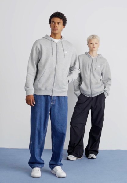 Dos modelos visten sudaderas con capucha y cremallera; una en gris claro y la otra también en gris. Las combinan con jeans de corte holgado y pantalones negros.