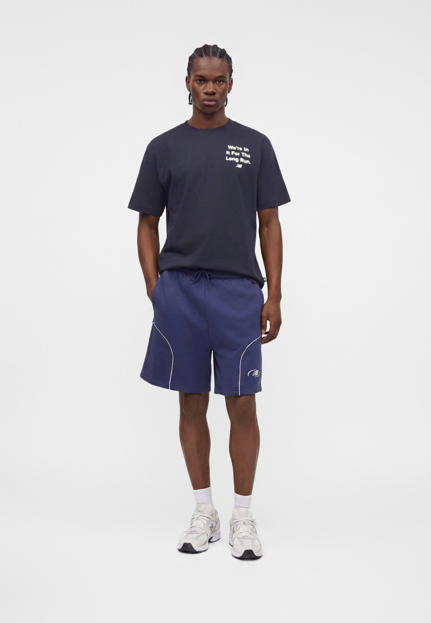 Hombre de pie con una camiseta negra con texto blanco y pantalones cortos azules con zapatillas blancas sobre un fondo blanco liso.