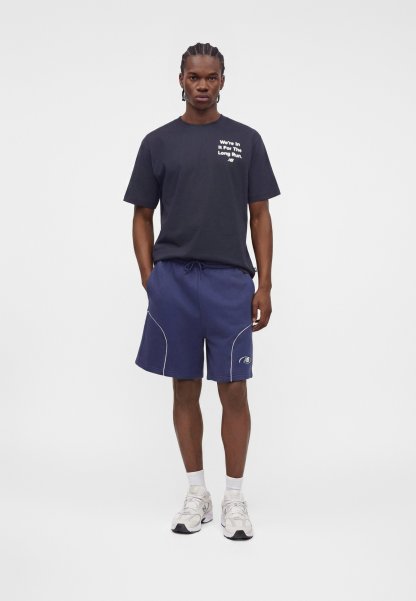 Hombre de pie con una camiseta negra con texto blanco y pantalones cortos azules con zapatillas blancas sobre un fondo blanco liso.