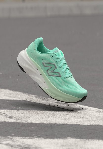 Zapatilla de running New Balance verde claro en el aire sobre el asfalto con partículas de suciedad y marcas de carretera blancas.