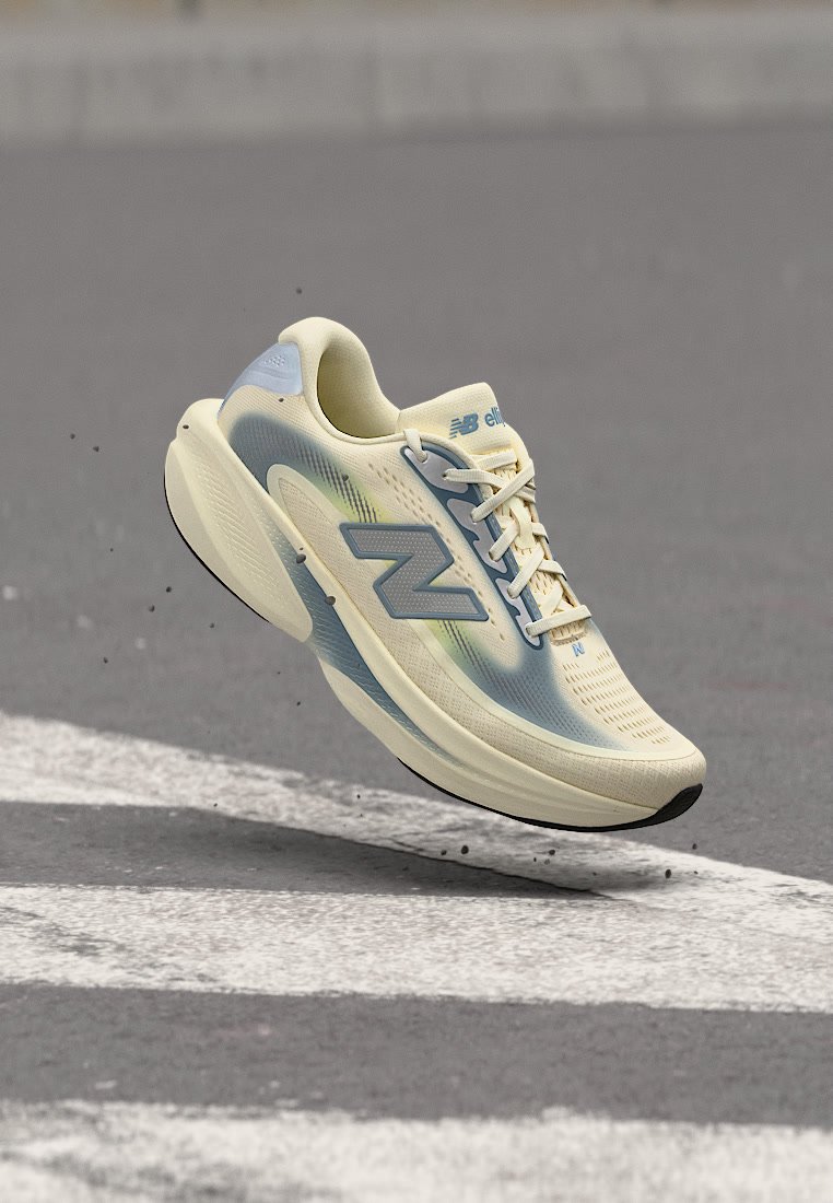 Zapatilla de running New Balance en beige claro y azul con cordones, flotando sobre asfalto con señales viales blancas.