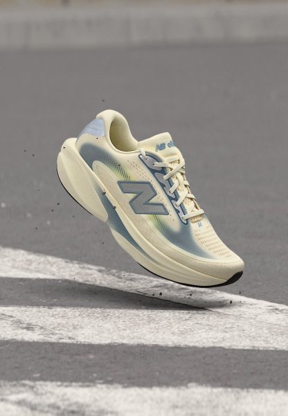 Zapatilla de running New Balance en beige claro y azul con cordones, flotando sobre asfalto con señales viales blancas.