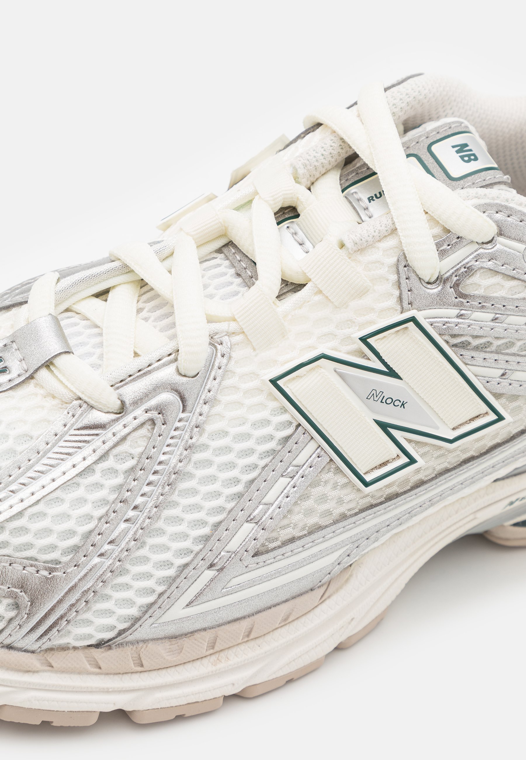 Zapatilla de running en malla blanca con detalles plateados. Presenta cordones en color crema y un logo prominente en verde y blanco. Suela texturizada para una mejor tracción.