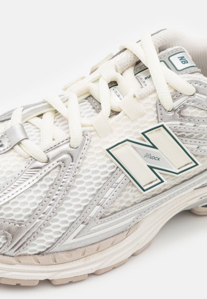 Zapatilla de running en malla blanca con detalles plateados. Presenta cordones en color crema y un logo prominente en verde y blanco. Suela texturizada para una mejor tracción.
