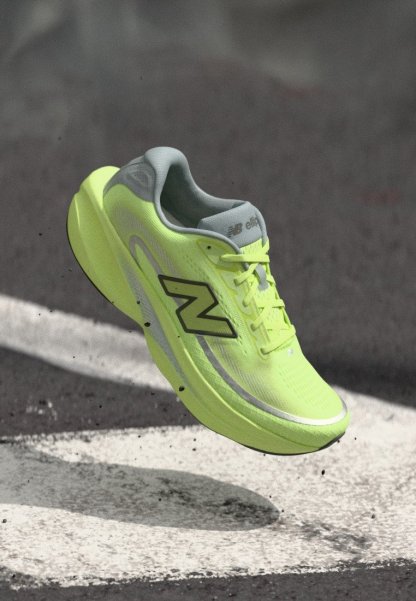 Zapatilla de correr New Balance de color amarillo brillante en el aire sobre asfalto con marcas blancas pintadas en la carretera, mostrando el logotipo en el lateral.
