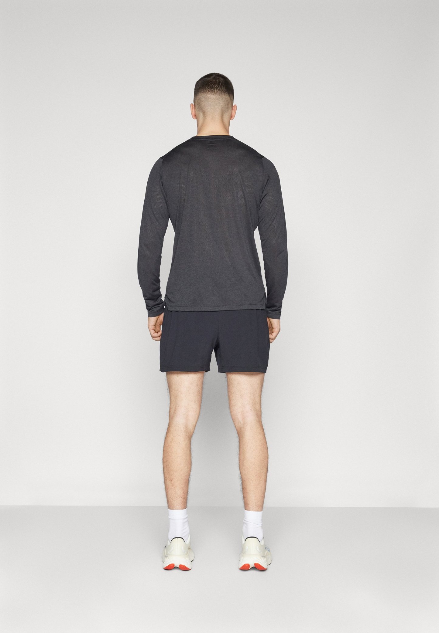 Camisa de manga larga gris combinada con pantalones cortos negros; vista desde la parte de atrás, con un diseño minimalista y un ajuste atlético. Zapatillas blancas visibles.
