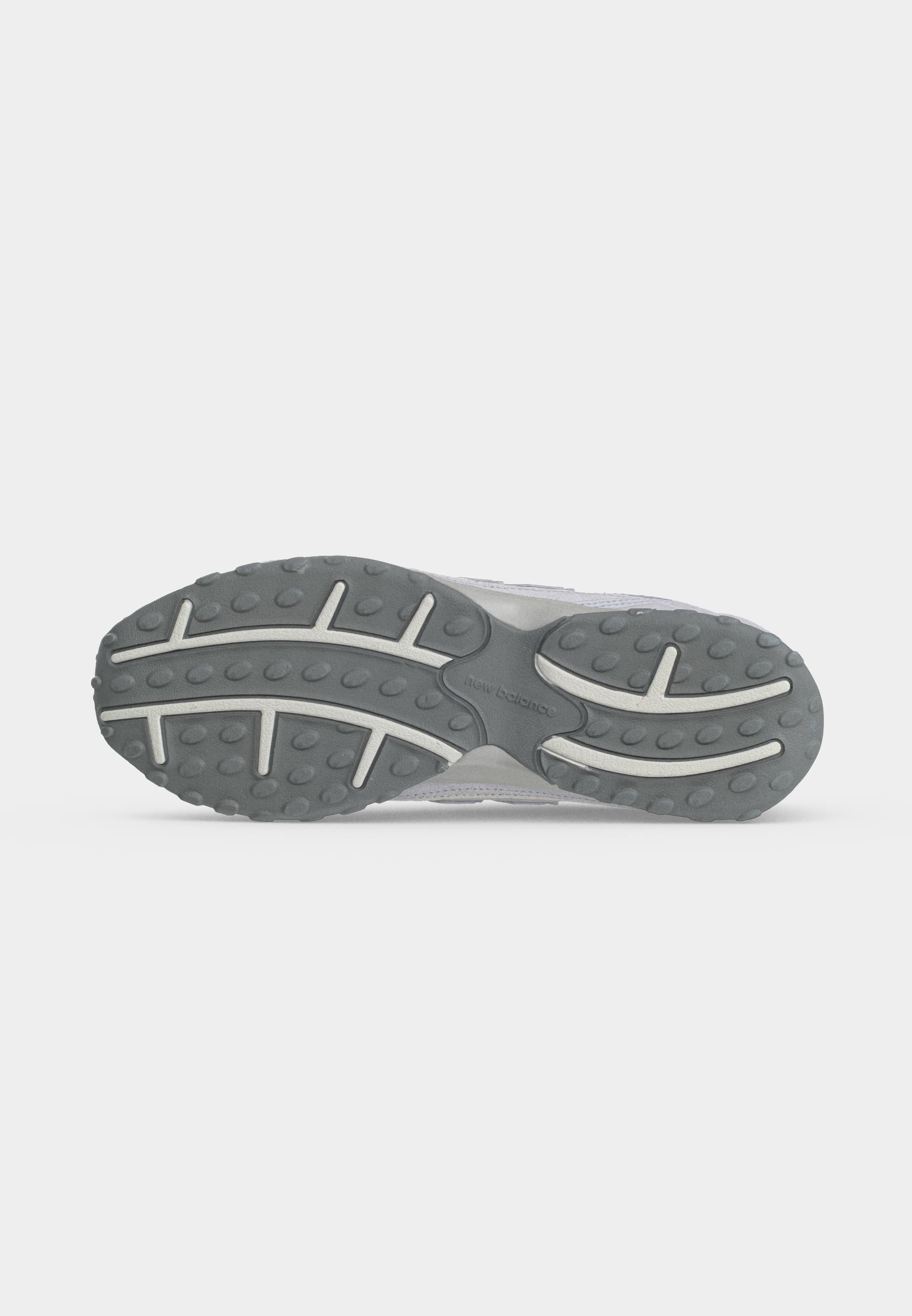 Suela de zapatilla gris y blanca con banda de rodadura de goma texturizada y logo de "New Balance" en el centro sobre un fondo claro y liso.