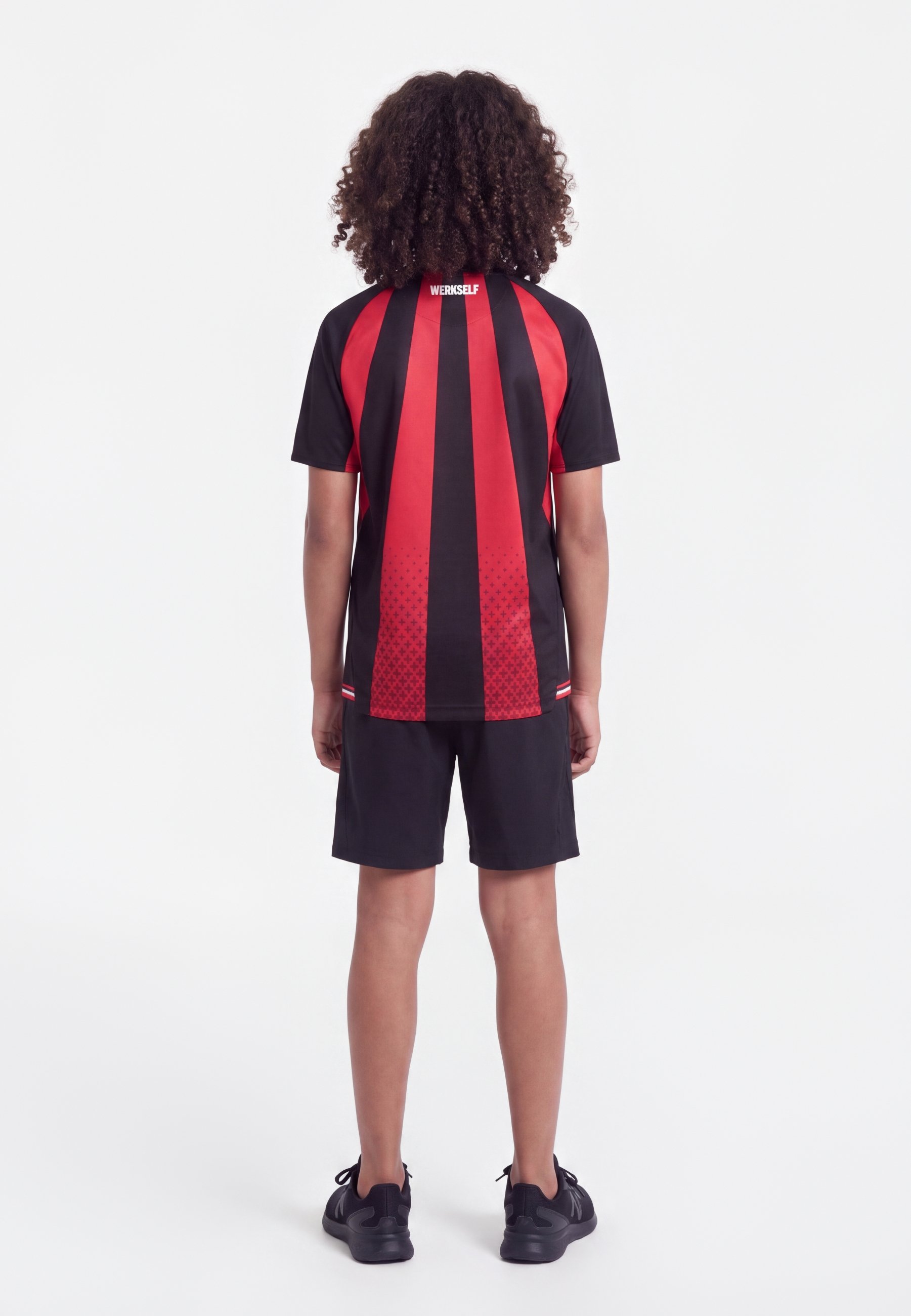 Niño con pelo rizado que lleva una camiseta deportiva de manga corta a rayas rojas y negras y pantalones cortos negros, de pie de espaldas sobre un fondo blanco.