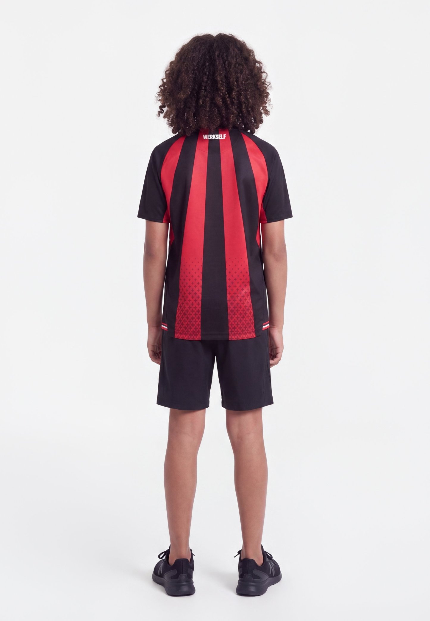 Niño con pelo rizado que lleva una camiseta deportiva de manga corta a rayas rojas y negras y pantalones cortos negros, de pie de espaldas sobre un fondo blanco.