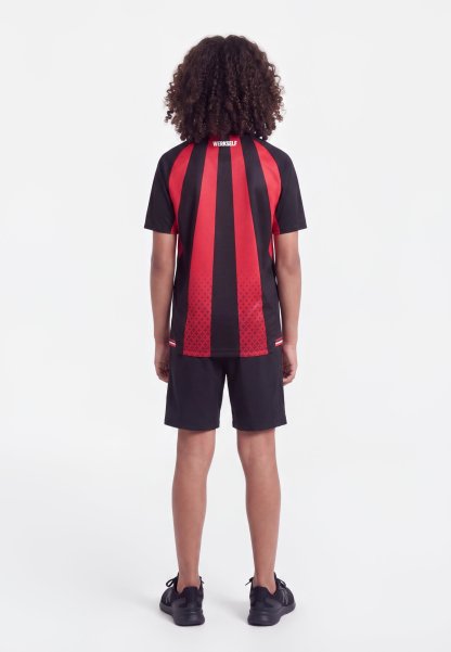Niño con pelo rizado que lleva una camiseta deportiva de manga corta a rayas rojas y negras y pantalones cortos negros, de pie de espaldas sobre un fondo blanco.