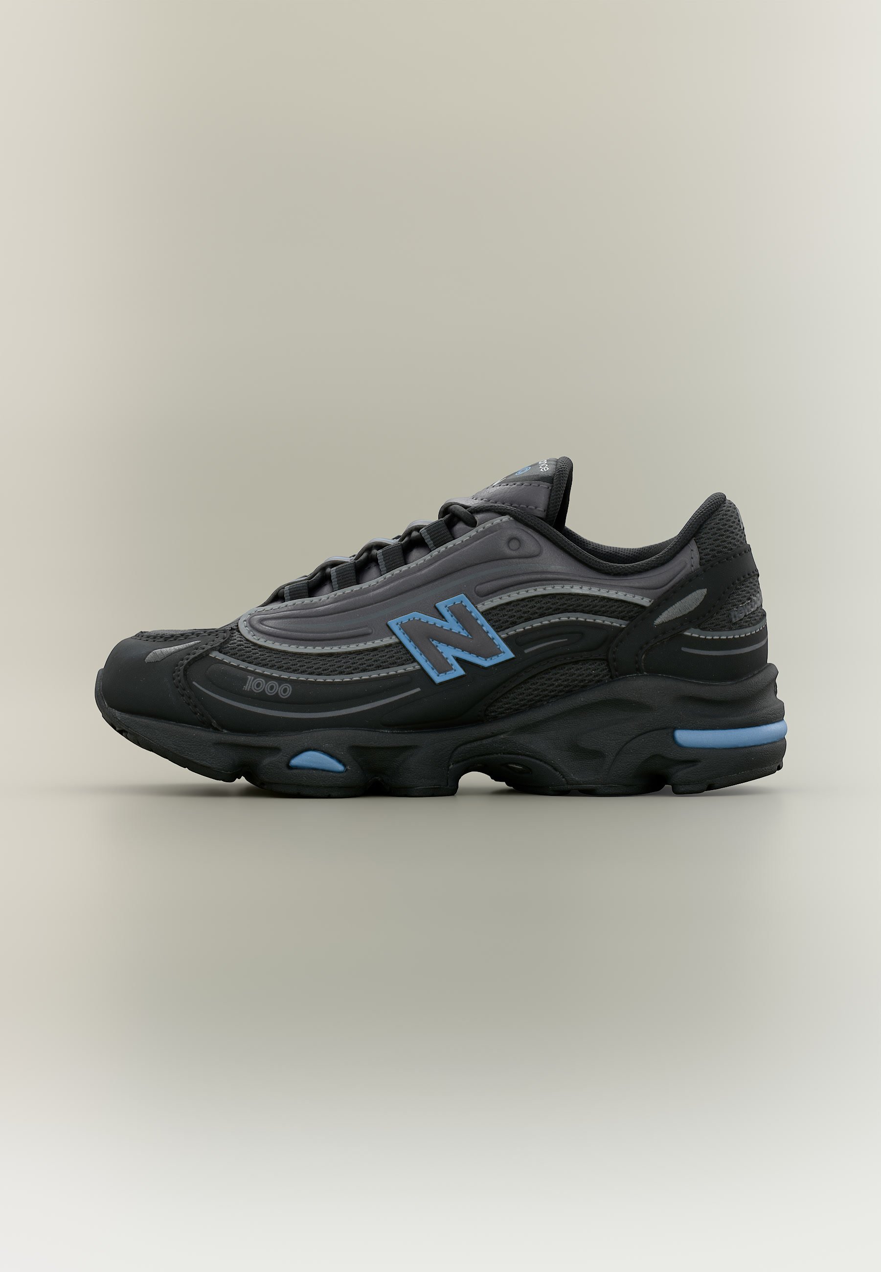 Zapatilla deportiva negra y gris con detalles azules y logo "N", diseño de caña baja y suela acolchada colocada sobre un fondo liso.