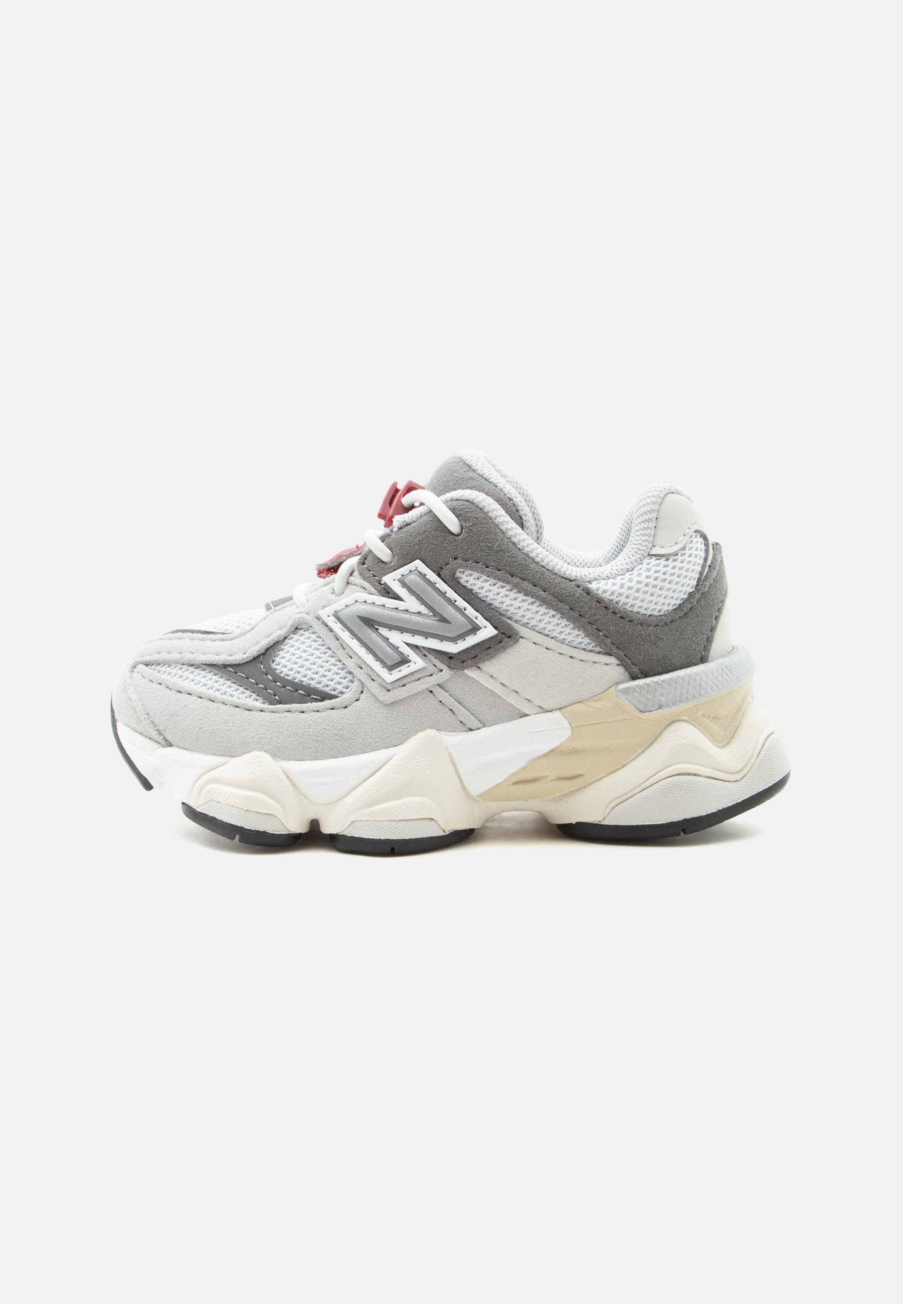 Zapatilla deportiva de malla y ante en gris y blanco con suela cremosa gruesa y cierre rojo para los cordones, vista de lado sobre fondo blanco.