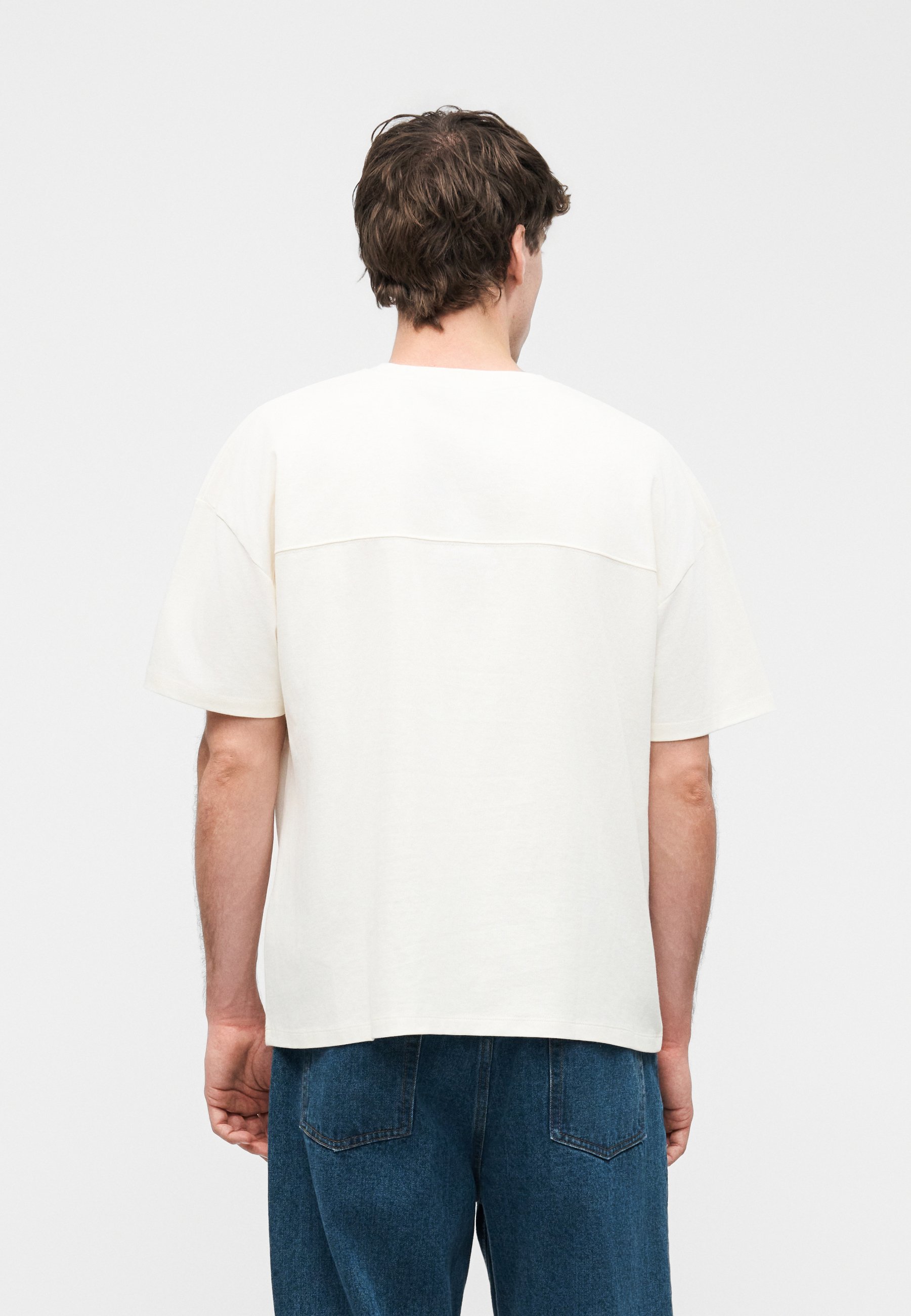 Hombre con cabello corto y marrón, vistiendo una camisa blanca de manga corta y suelta y vaqueros azules, de pie de espaldas sobre un fondo liso.