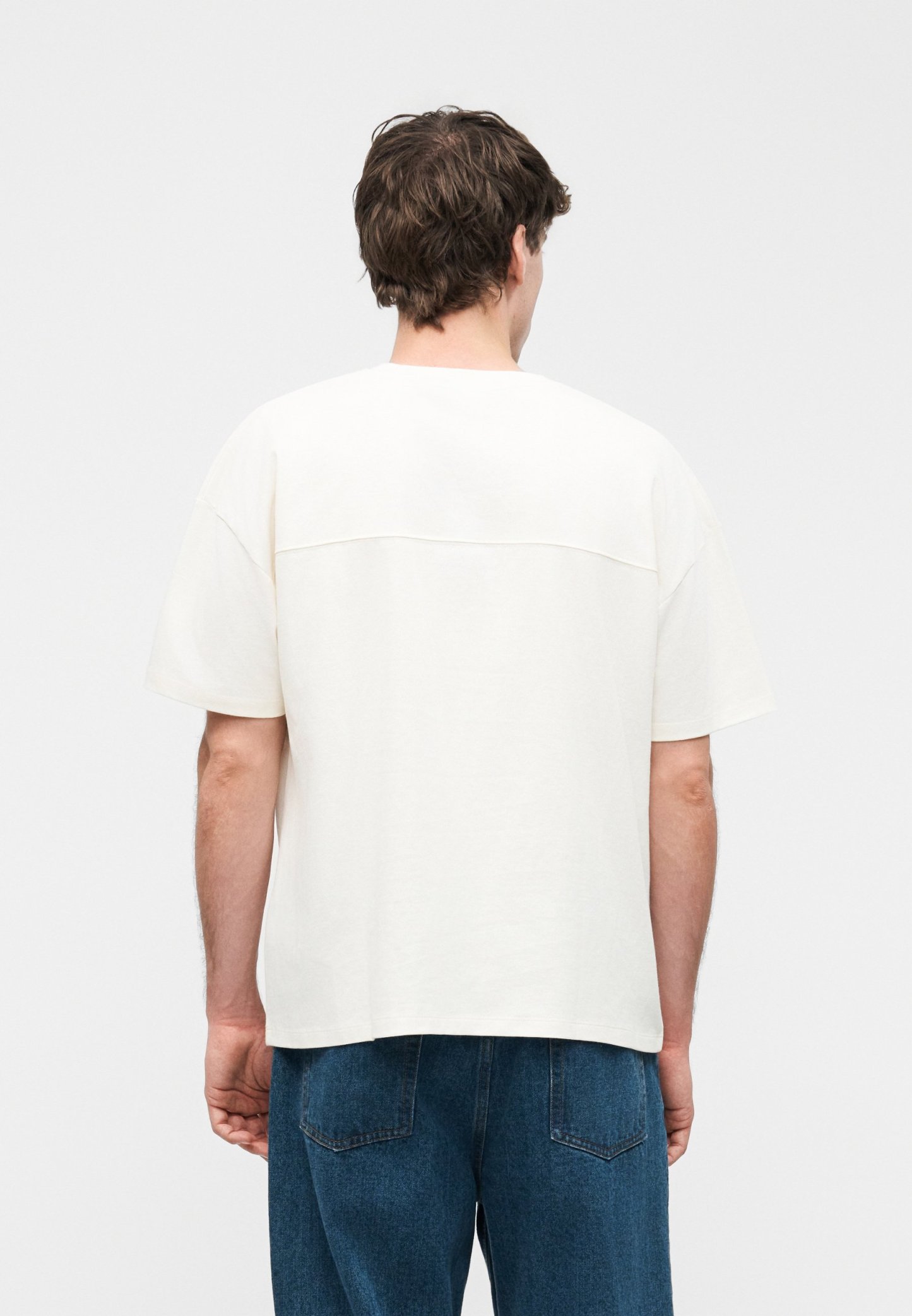 Hombre con cabello corto y marrón, vistiendo una camisa blanca de manga corta y suelta y vaqueros azules, de pie de espaldas sobre un fondo liso.