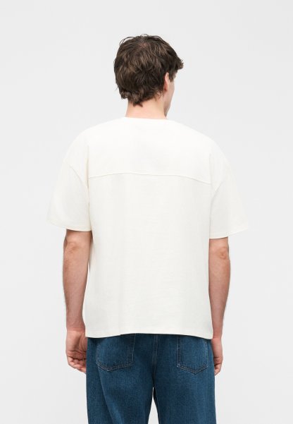 Hombre con cabello corto y marrón, vistiendo una camisa blanca de manga corta y suelta y vaqueros azules, de pie de espaldas sobre un fondo liso.