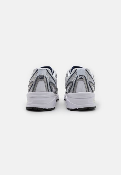 Zapatillas deportivas blancas y grises con parte superior de malla, diseño curvado y suela texturizada. Presenta un pequeño logo en la parte trasera y un talón contorneado.