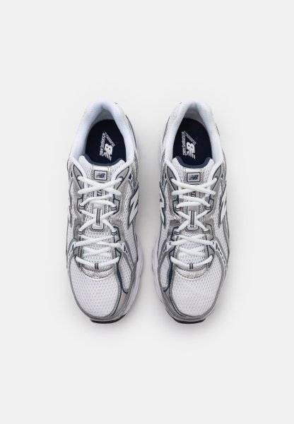 Zapatos deportivos blancos con parte superior de malla, acentos en gris, cierre de cordones tradicional y suela acolchada. Presenta un logo en la lengüeta y en el lateral.