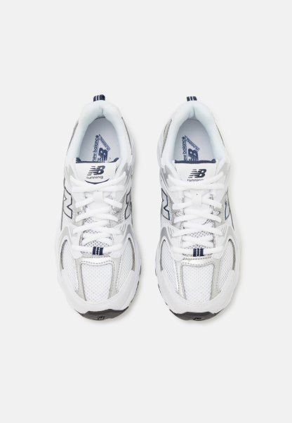 Zapatillas deportivas blancas con parte superior de malla y sintético, con una suela gris, acentos en azul marino y cordones en contraste. Se pueden ver dos logotipos de "N".