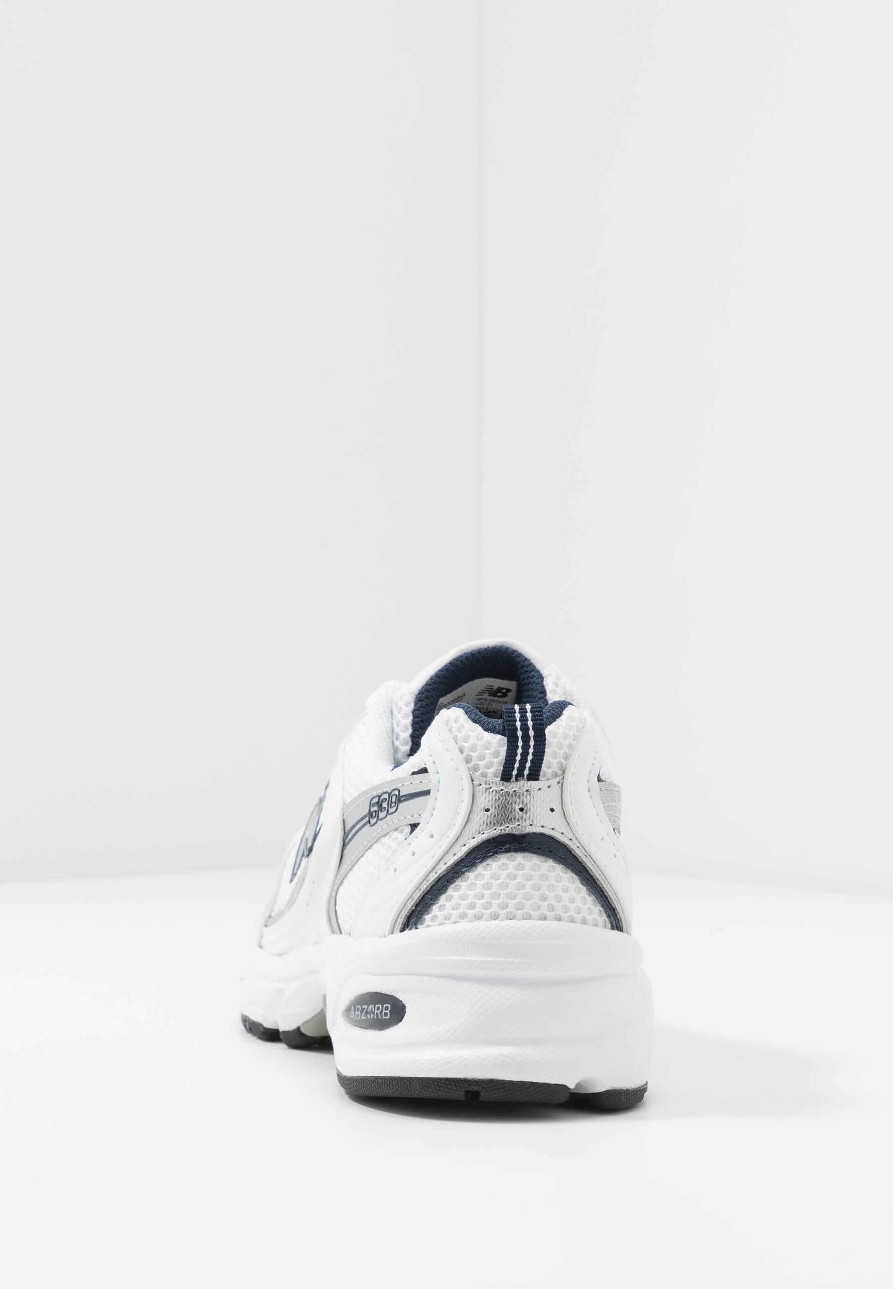 Zapatilla deportiva blanca con parte superior de malla, acentos grises, cuello acolchado y suela de goma. Presenta una franja gris y el logo de la marca en el costado.