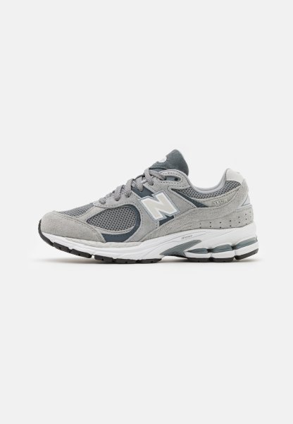 Zapatilla gris con materiales de malla y gamuza, que presenta un logo blanco, un cuello acolchado y una suela de goma texturizada. Detalles en un gris más oscuro.