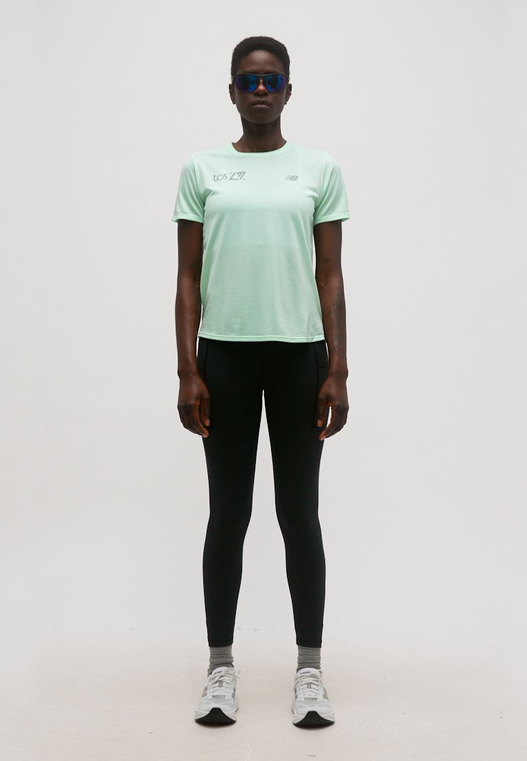 Persona vistiendo una camiseta deportiva de manga corta color verde menta, leggings negros, calcetines grises, zapatillas blancas y gafas deportivas azul reflectantes, de pie y erguida.