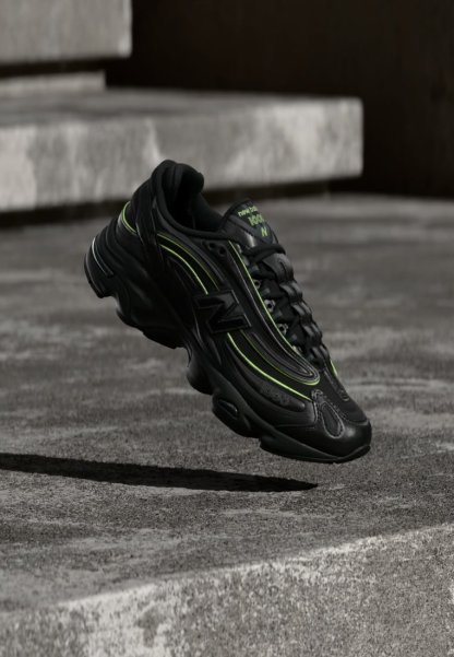 Zapatilla negra New Balance con detalles verdes flotando sobre escalones de hormigón gris en un entorno exterior.
