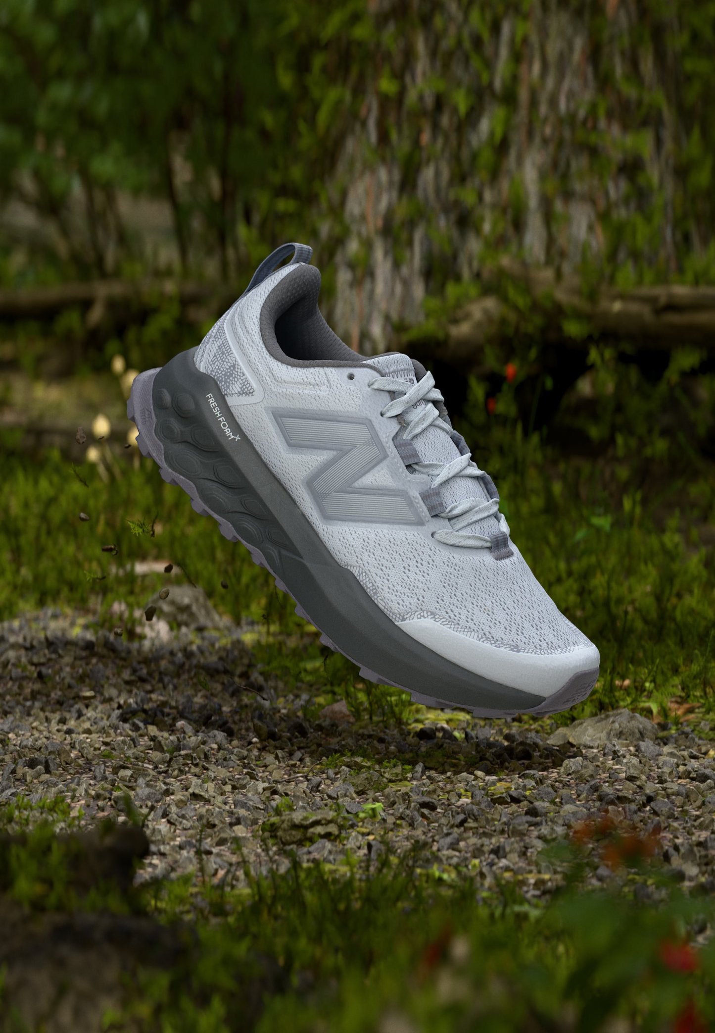 Zapatilla de running New Balance gris clara con suela gruesa gris suspendida sobre un camino de grava en un entorno natural al aire libre.