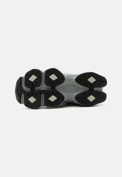 Suela de zapatilla New Balance gris con bandas de goma negras y patrones de tracción en forma de diamante de color verde claro sobre un fondo blanco.