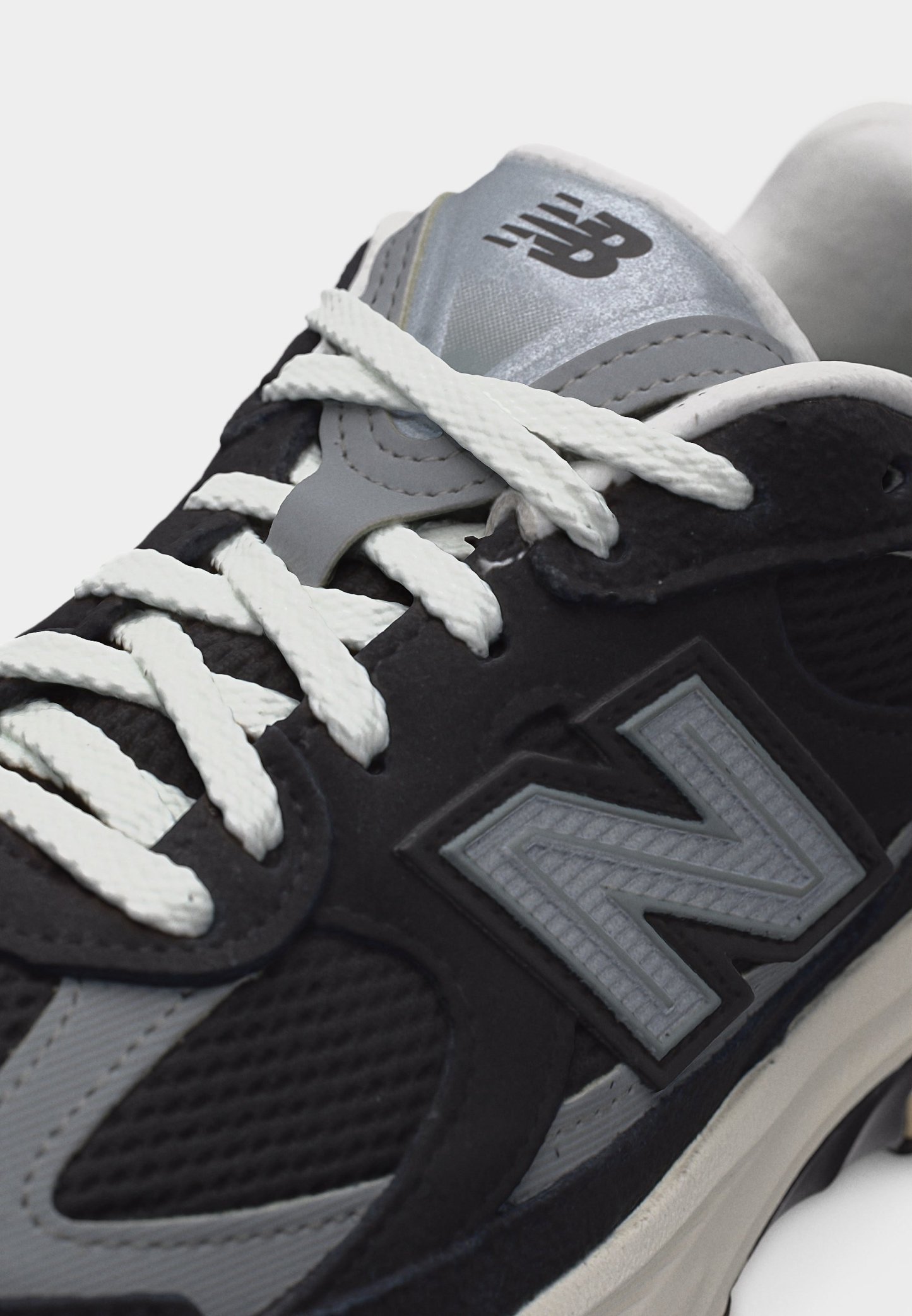 Zapatilla New Balance en negro y gris con cordones blancos y logo en el lateral y la lengüeta, mostrada en un ángulo de primer plano.