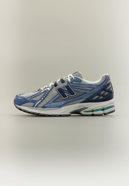 Zapatilla deportiva New Balance en gris y azul con cordones blancos, suela acolchada y parte superior de tela de malla vista de lado sobre fondo neutro.