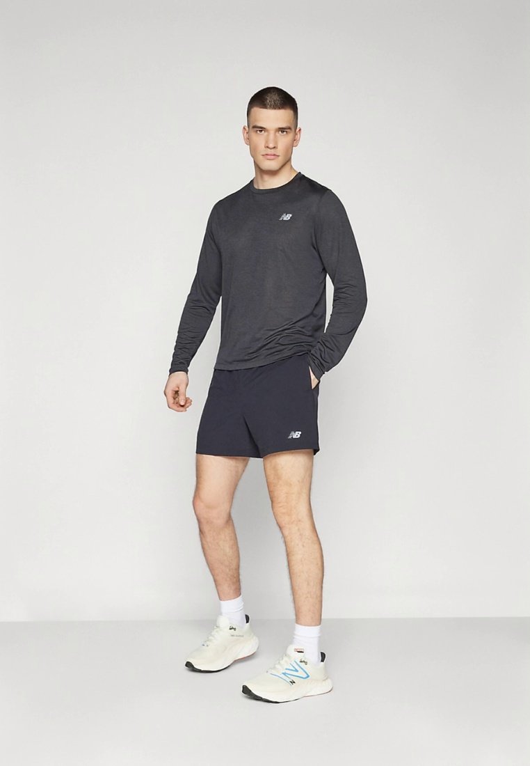 Camiseta deportiva negra de manga larga, pantalones cortos azul marino, zapatillas blancas con detalles en azul. Modelo de pie centrado sobre un fondo liso.