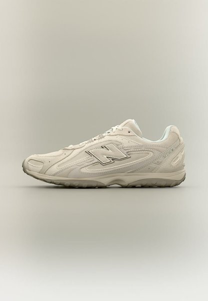 Zapatilla blanca New Balance 2011 con parte superior de ante y malla, suela gris y diseño de cordones, mostrada desde el lado exterior sobre un fondo neutro.