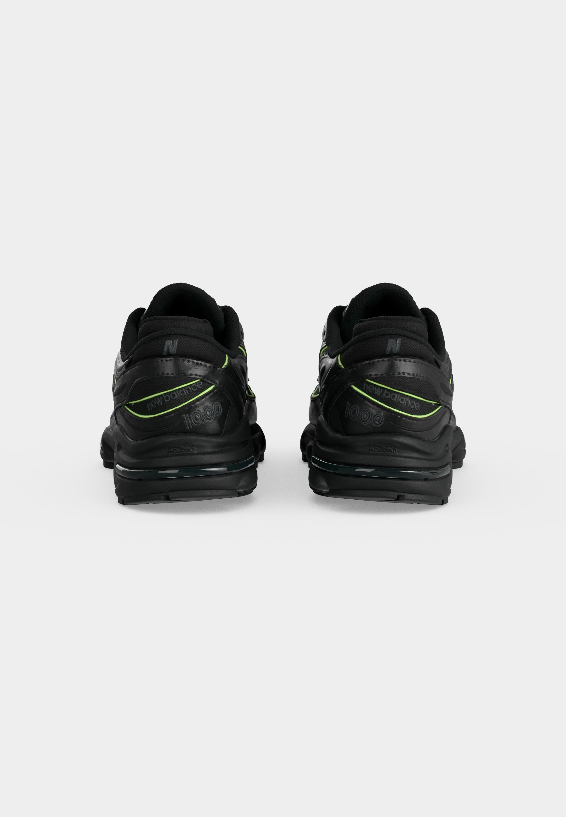 Par de zapatillas negras New Balance 1000 con detalles en verde neón, mostradas desde atrás sobre un fondo liso gris claro.