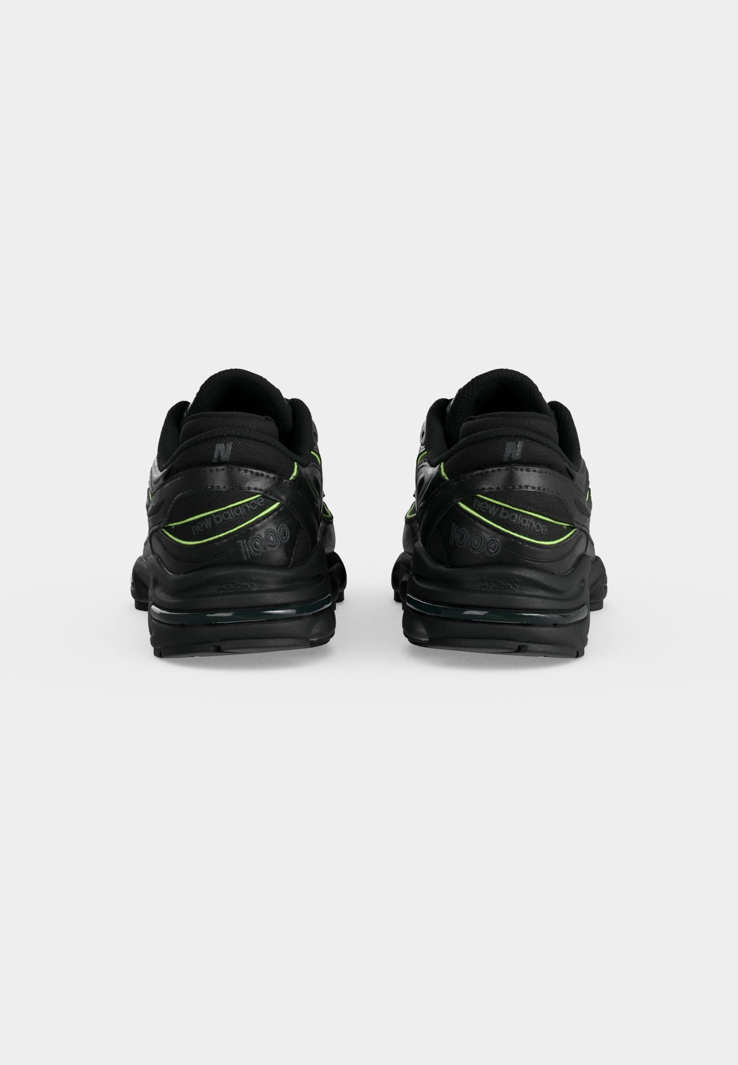 Par de zapatillas negras New Balance 1000 con detalles en verde neón, mostradas desde atrás sobre un fondo liso gris claro.