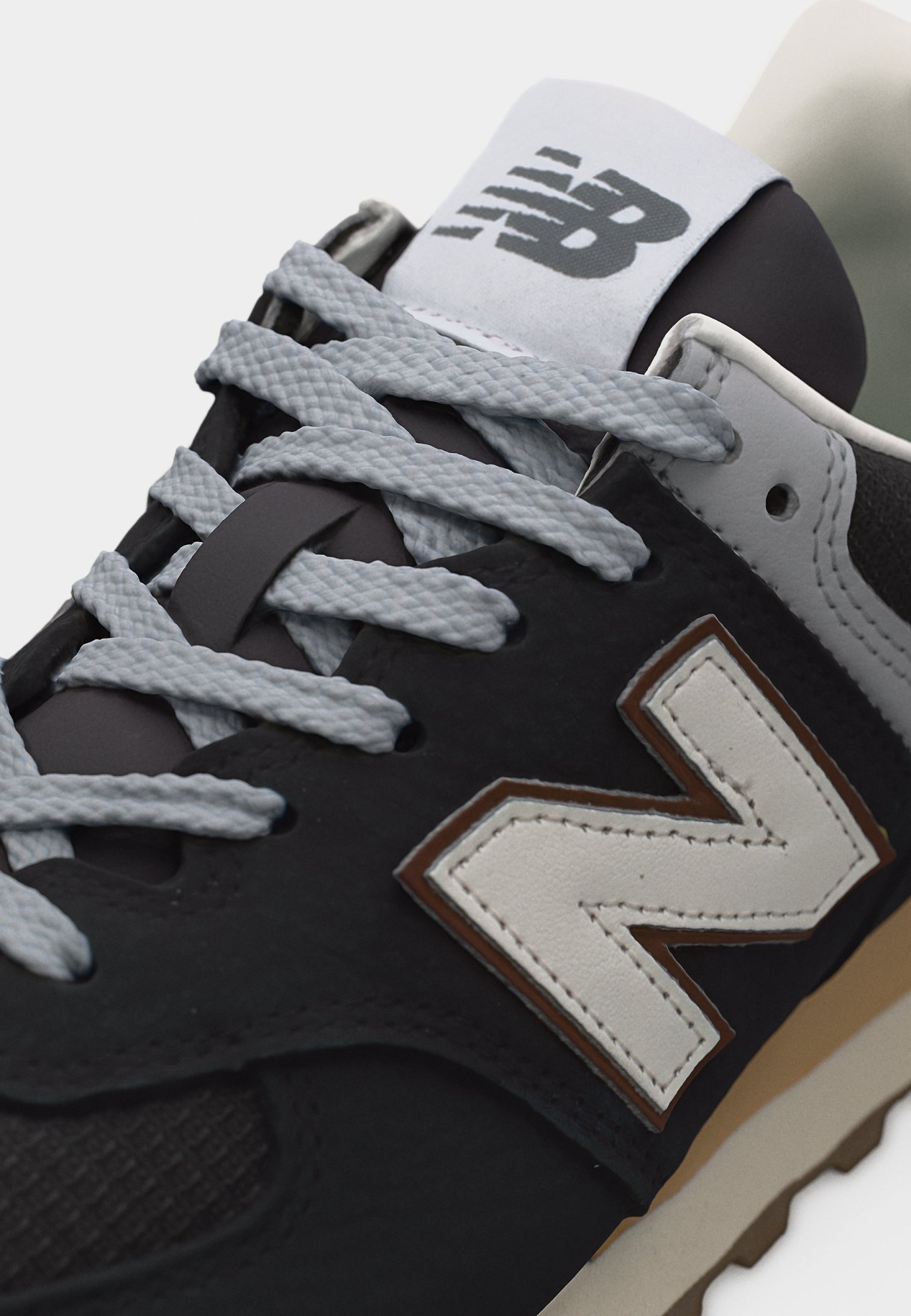 Zapatilla New Balance en blanco y negro con cordones grises, logotipo blanco "N" delineado en marrón y suela beige visible en el borde inferior.