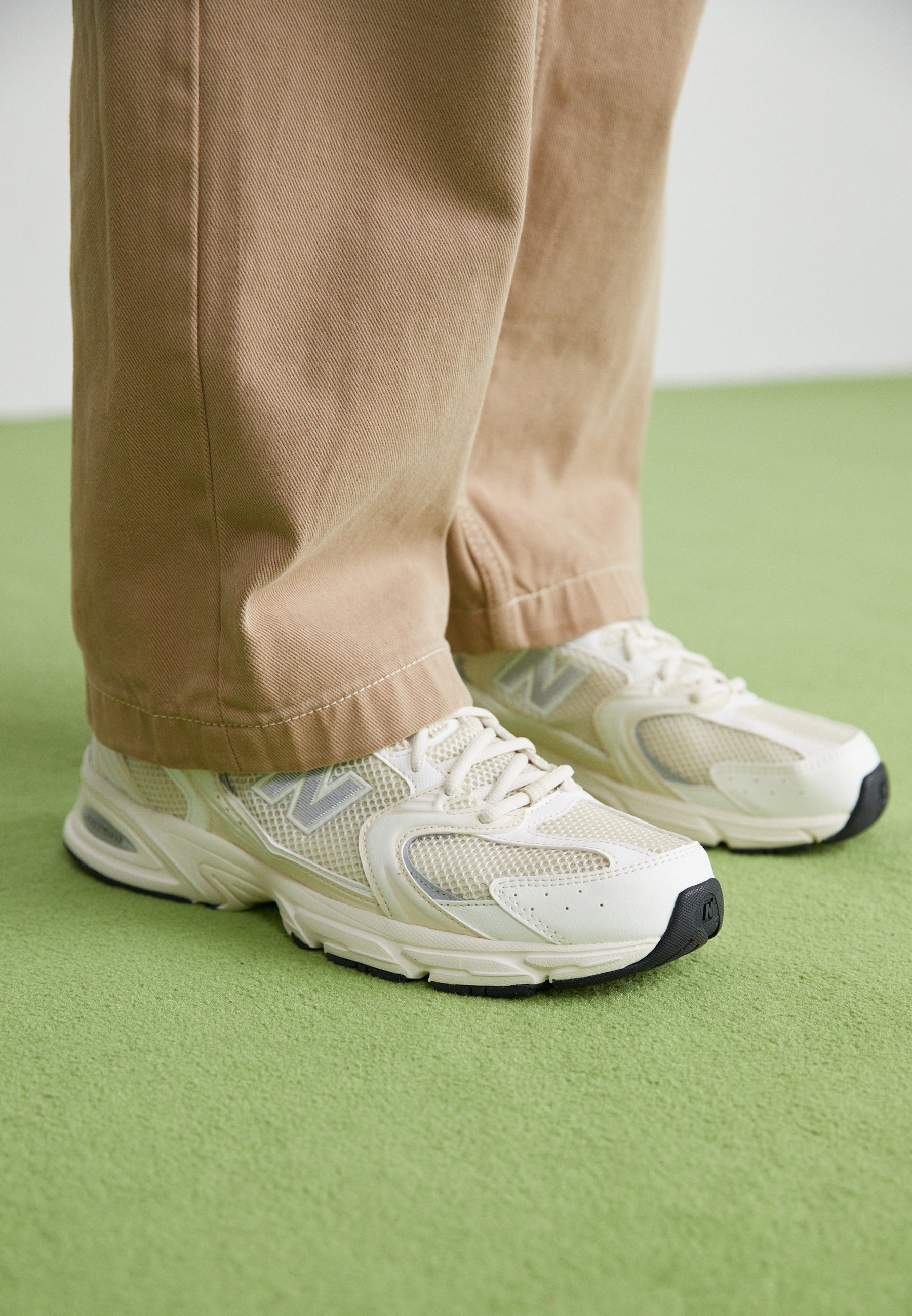 Zapatillas para correr de malla blanca y beige, usadas con pantalones beige sobre un suelo verde texturizado, mostrando vistas frontal y lateral de ambos pies.
