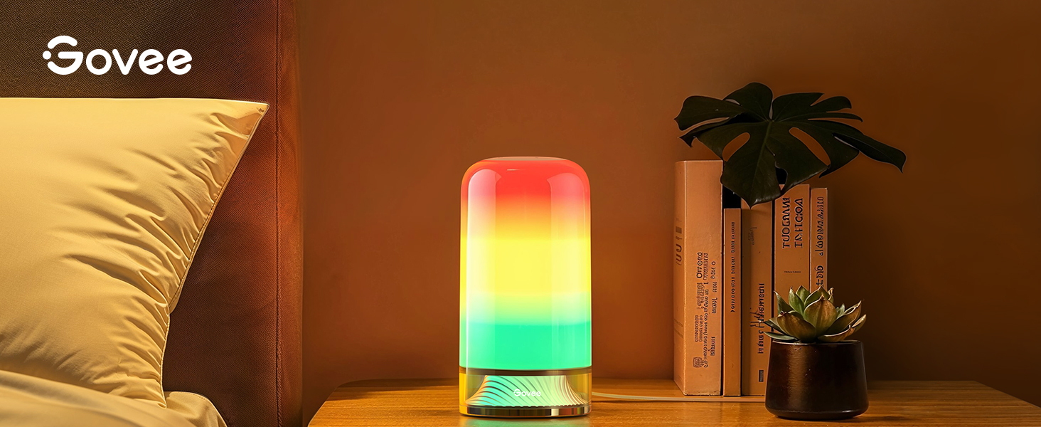 Lampe d'ambiance cylindrique à couleur changeante diffusant une lumière dégradée rouge, jaune et verte, positionnée sur une table de chevet avec des plantes décoratives à proximité