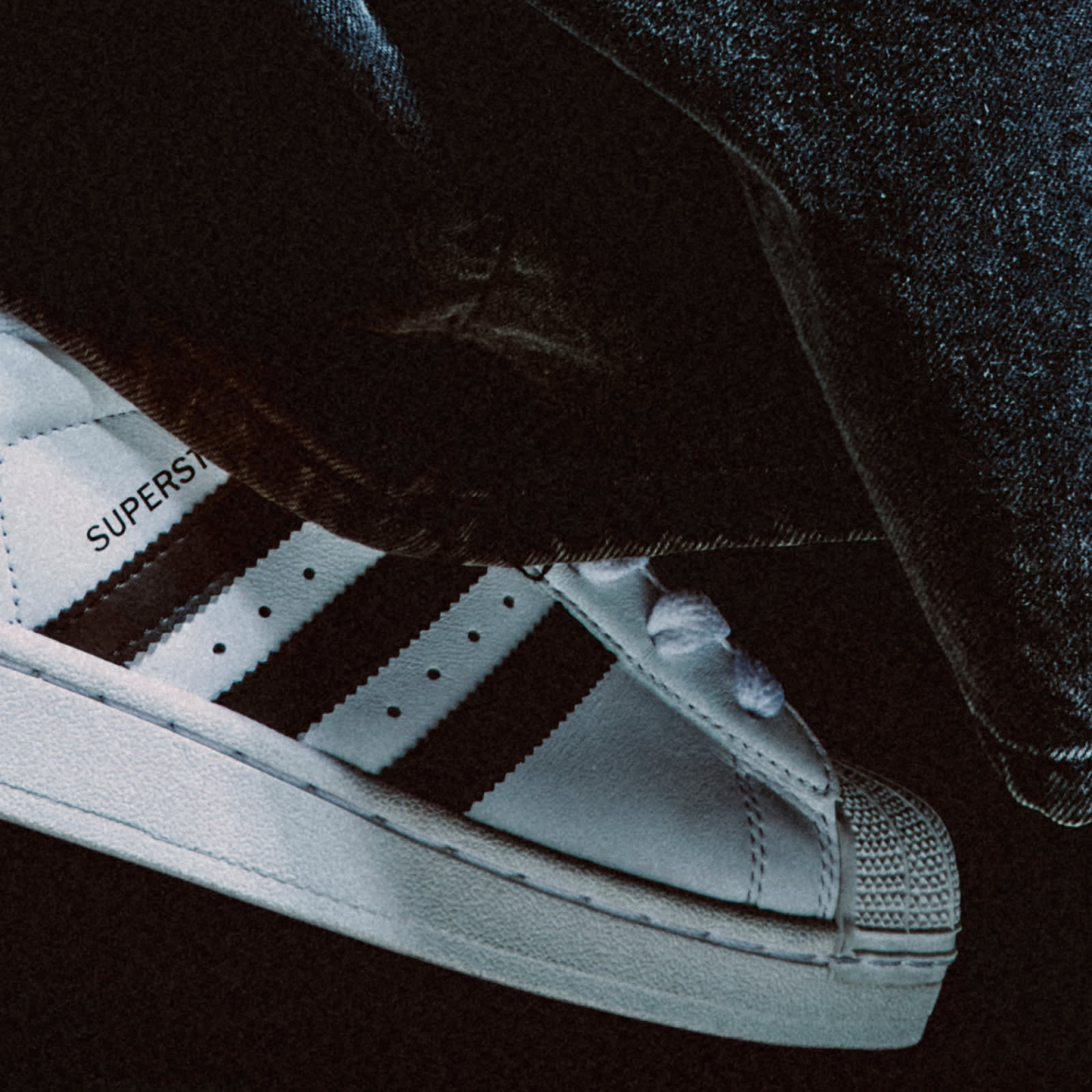 A close-up image of a person wearing a white Adthtidas Superstar sneaker with black stripes and grey denim jeans standing on a concrete floor.