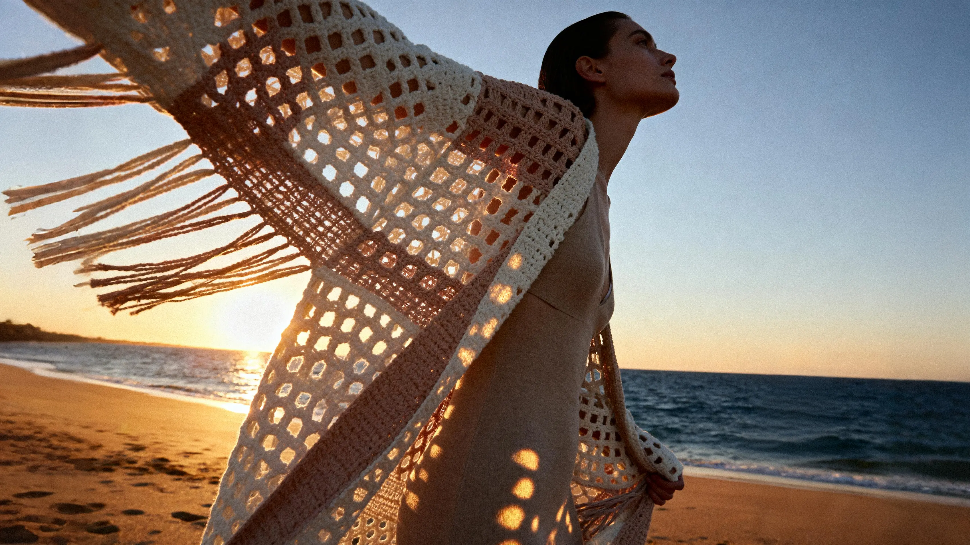 {"default":"Crochet clothing collection banner featuring a model wearing a beige open-knit cardigan by the sea at sunset"}
