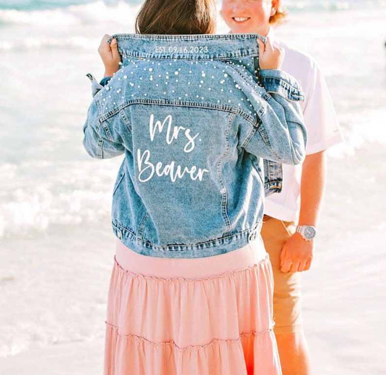 May include: A blue denim jacket with pearl embellishments on the back. The jacket has white text on the back that says 'Mrs. Beaver' and 'Est. 09.16.2023'.