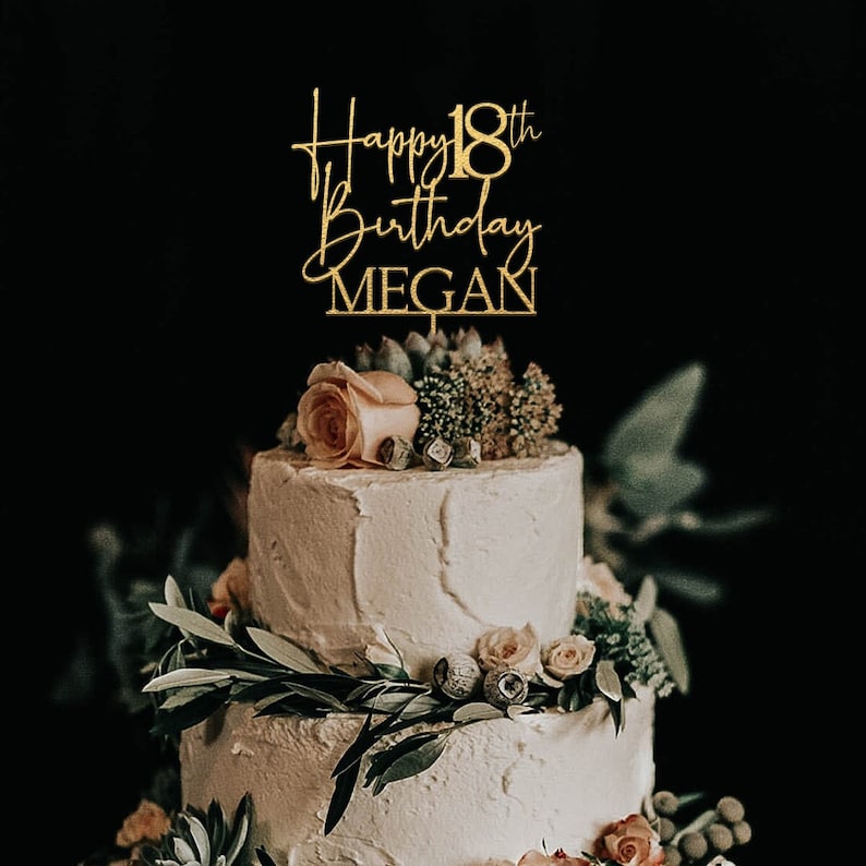May include: A two-tiered white cake with gold glitter cake topper that reads 'Happy 18th Birthday MEGAN' and is decorated with greenery and small pink roses.